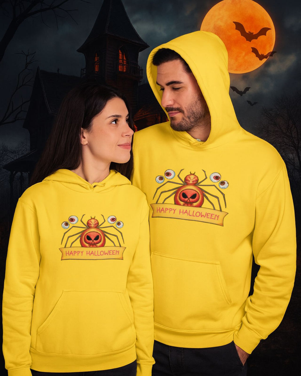 A couple wearing a yellow sweatshirt with the phrase "Happy Halloween" printed on it and an orange spider with orange eyes on it, the boy has the hood up and looks at her and she looks at him, in the background you can see an image of a haunted house with lights inside and a giant orange moon over which bats fly, underneath they are wearing jeans